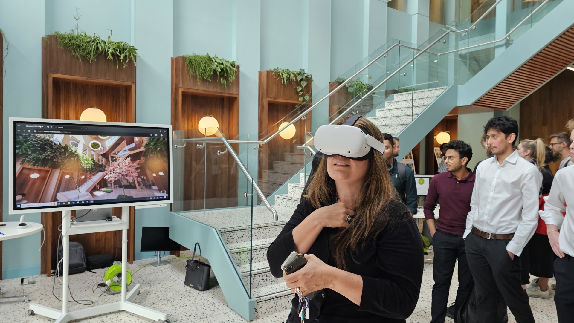 A woman wears a VR headset and she is stood in an atrium. Behind her is a screen with atrium covered in plants and trees